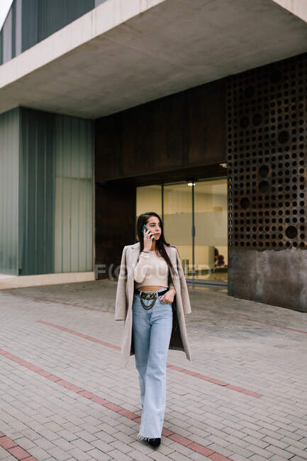 Empresária na moda em roupas elegantes andando na cidade e falando no telefone celular enquanto olha para longe — Fotografia de Stock