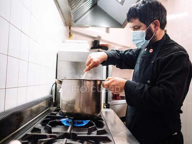 Side view of professional male cook in mask stirring dish in saucepan while cooking on stove in kitchen of restaurant — Stock Photo