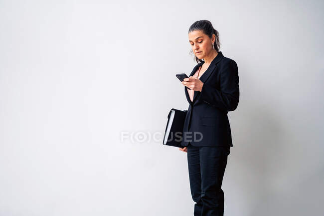 Серьёзная предпринимательница средних лет с хвостиком в чёрном костюме и текстовыми сообщениями на мобильном телефоне, стоя на белом фоне с папкой — стоковое фото