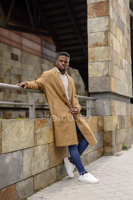 Full body of African American male wearing trendy coat leaning stone wall of building in city and looking away — Stock Photo