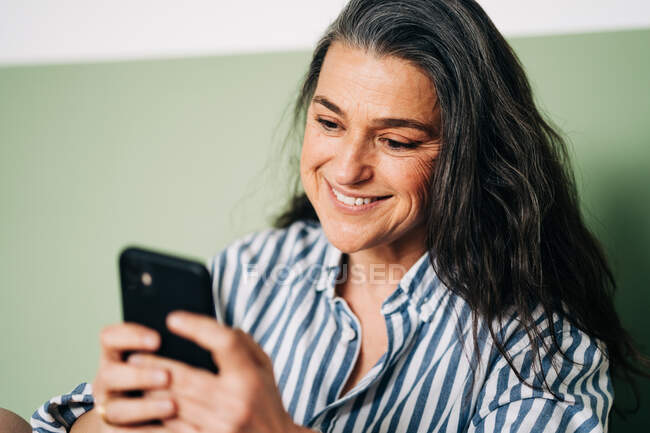 Позитивна жінка середнього віку з довгим темним волоссям в повсякденному одязі посміхається під час розмови на мобільному телефоні, сидячи на ліжку вдома. — стокове фото