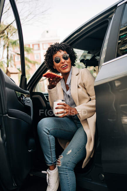Happy African American female in fashionable sunglasses with thermal mug and recording audio message on smartphone while smiling and leaving silver prestige automobile — Stock Photo