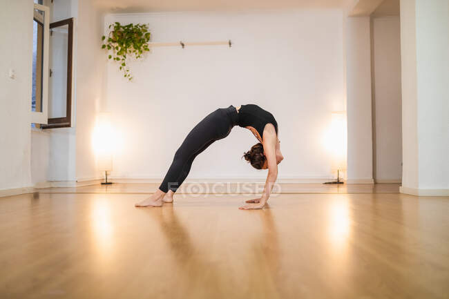 Vista lateral de una hembra flexible irreconocible en ropa deportiva de pie en Urdhva Dhanurasana posando mientras practica yoga en suelo brillante - foto de stock