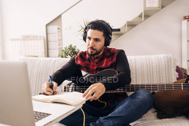 Adult male guitarist in headphones with electric guitar taking notes in notebook while composing music against netbook on sofa at home — Stock Photo