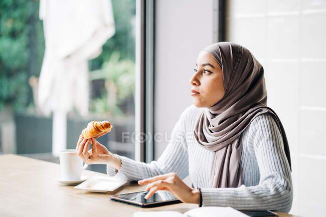 Этнические женщины в хиджаб серфинг Интернет на планшете, сидя за столом с круассаном и чашкой кофе в кафе и наслаждаясь выходные — стоковое фото