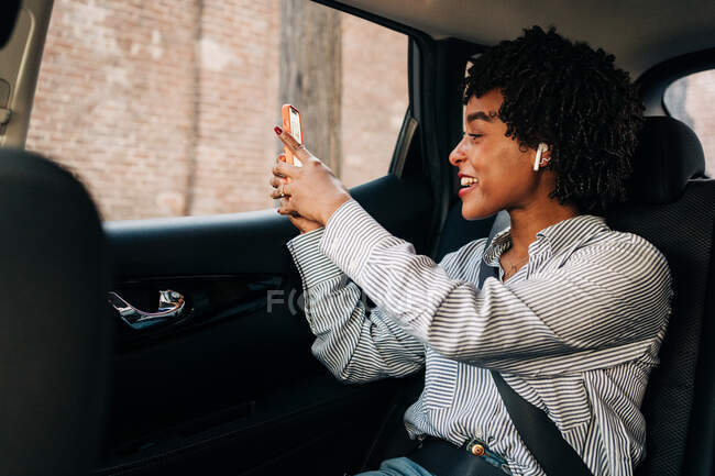 Вид сбоку жизнерадостной афроамериканки, улыбающейся и разговаривающей в видеочате в мобильном телефоне в современном автомобиле — стоковое фото