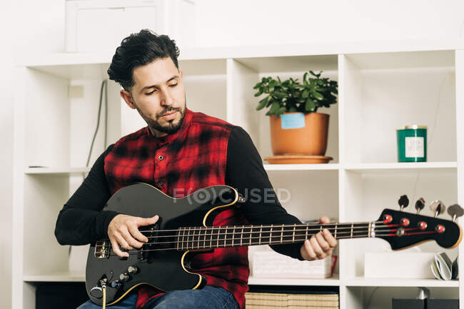 Musicien barbu professionnel adulte jouant de la guitare basse pendant la répétition contre les étagères à la maison — Photo de stock