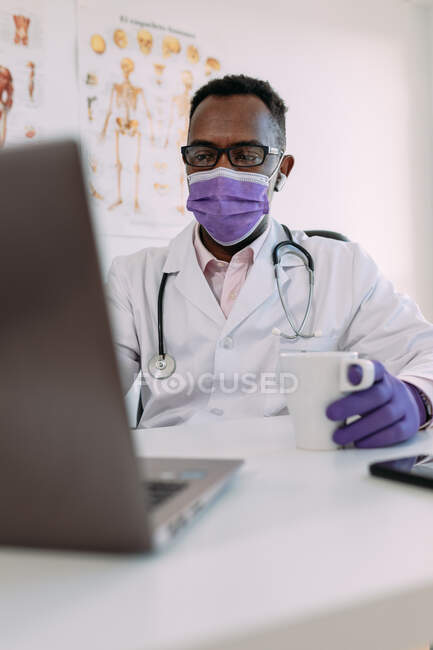 Ireconhecível concentrado Africano americano médico masculino em roupão médico e máscara beber café e trabalhar no laptop na clínica moderna — Fotografia de Stock