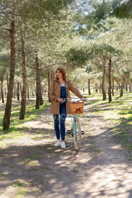 Повне тіло весела молода жінка посміхається і дивиться далеко біля старого велосипеда з дерев'яним плетеним кошиком — стокове фото
