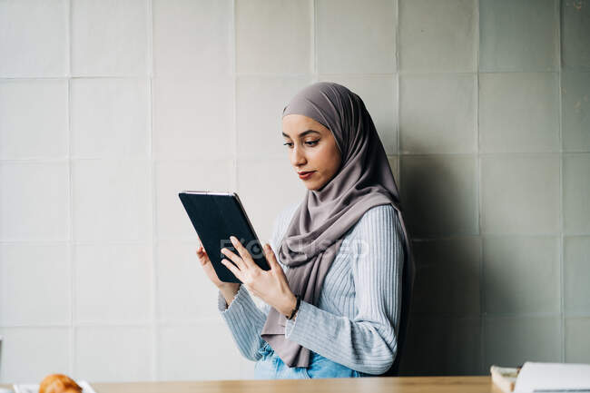 Vista laterale della freelance musulmana che indossa hijab tradizionale in piedi nel caffè e tablet di navigazione mentre lavora al progetto in remoto — Foto stock