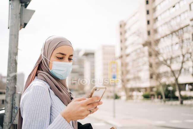 Side view of ethnic female wearing headscarf and protective mask standing on street in city on sunny day and looking at screen of her mobile — Stock Photo