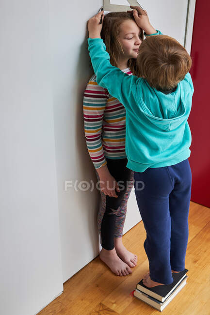 Brother helping sister with measuring her height with ruler and pencil near wall — Stock Photo