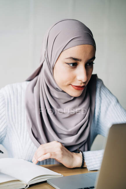 Portrait of ethnic female freelancer in hijab using laptop while sitting at table in cafe and working remotely — Stock Photo