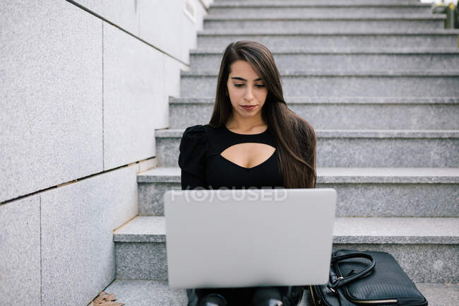 Femme entrepreneure concentrée assise sur des marches en pierre dans la ville et naviguant sur le netbook tout en travaillant sur le projet en ligne — Photo de stock