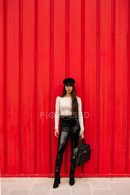 Female entrepreneur wearing trendy clothes standing on the street on background of red wall and looking at camera — Stock Photo