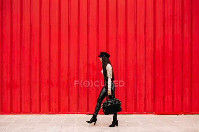 Vista lateral de empresária vestindo roupas da moda andando ao longo da rua no fundo da parede vermelha e olhando para longe — Fotografia de Stock