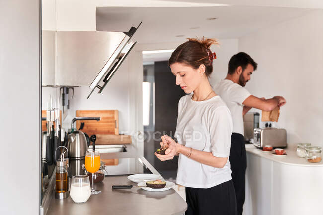 Vista lateral da calma jovem mulher em t-shirt branca preparando café da manhã saudável com abacate enquanto está à mesa na cozinha perto do namorado preparando algumas torradas — Fotografia de Stock
