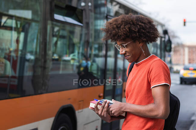 Vista laterale di moda giovane studente nero maschio con i capelli afro in abiti casual e occhiali che tengono libri mentre in piedi sulla strada della città vicino a autobus moderno — Foto stock