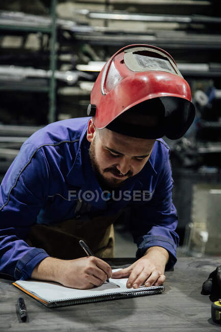 Serious bearded male worker in hardhat and overall taking notes in notebook with pen on table near metal constructions in light garage — Stock Photo