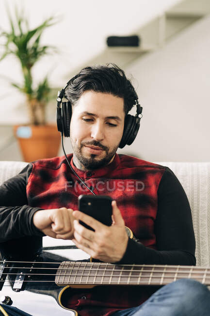 Adulto barbudo músico masculino com baixo guitarra em fones de ouvido mensagens de texto no celular na sala de estar — Fotografia de Stock