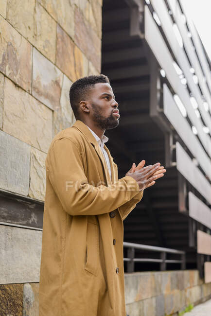 Vue de côté homme afro-américain portant manteau à la mode debout près du mur de pierre du bâtiment en ville et mettre les mains ensemble — Photo de stock