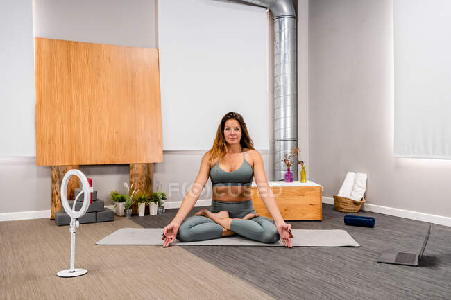 Voller Körper einer konzentrierten jungen Frau in aktiver Kleidung, die in Lotus-Pose sitzt, in die Kamera blickt und während der Yoga-Sitzung zu Hause in der Nähe des Smartphones meditiert, das auf einem Stativ steht — Stockfoto