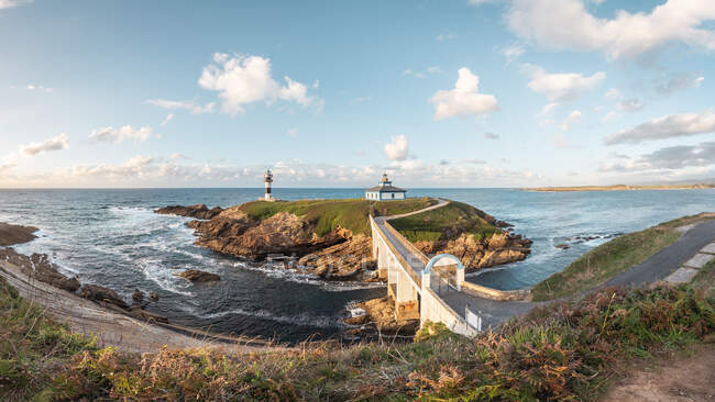 Paisagem espetacular de ponte que leva a ilha rochosa coberta com grama verde com farol colocado em oceano ondulado em Faro Illa Pancha, na Galiza, na Espanha, durante o dia — Fotografia de Stock