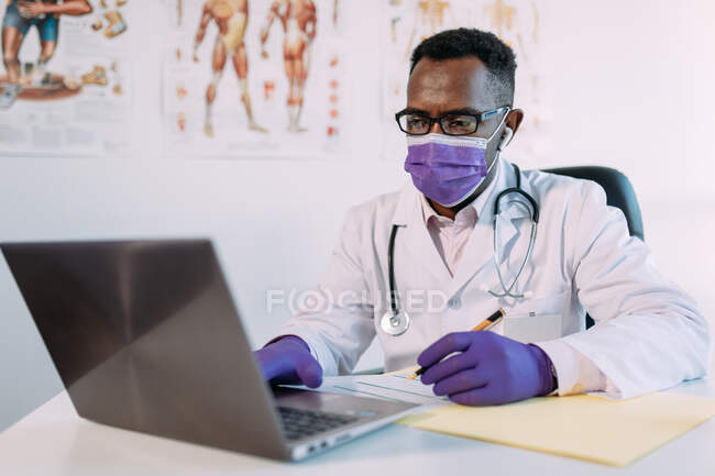 Medico afroamericano in occhiali che lavora con il paziente online su netbook mentre scrive nella cartella del paziente a tavola in ospedale — Foto stock