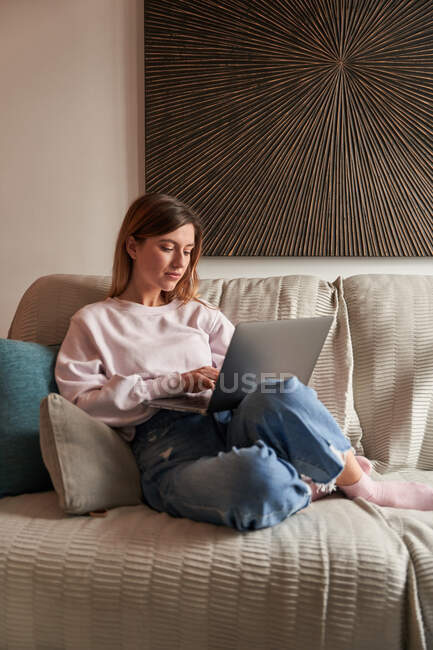 Ganzkörper friedliche junge Freiberuflerin in lässiger Kleidung auf gemütlichem Sofa sitzend und ferngesteuert am Laptop arbeitend — Stockfoto