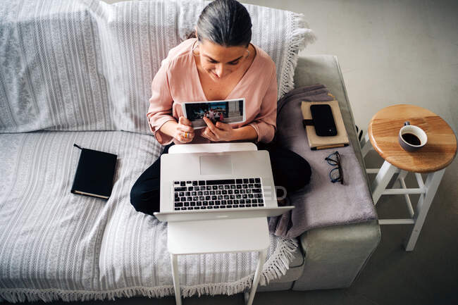 Von oben von einer Frau, die auf dem Sofa sitzt und ein Bild zeigt, während sie im heimischen Wohnzimmer einen Videochat per Laptop führt — Stockfoto