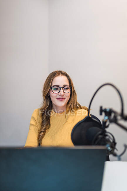 Mujer positiva navegando netbook moderno mientras está sentada en la mesa con monitores y micrófono durante el trabajo en el estudio de difusión con equipos especiales - foto de stock