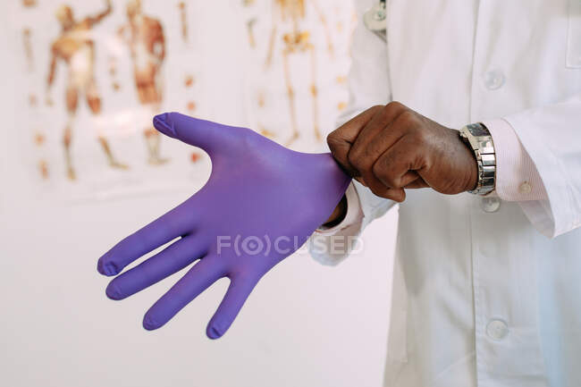 Crop anonymous African American male putting medical glove on hand on blurred background of modern hospital — Stock Photo