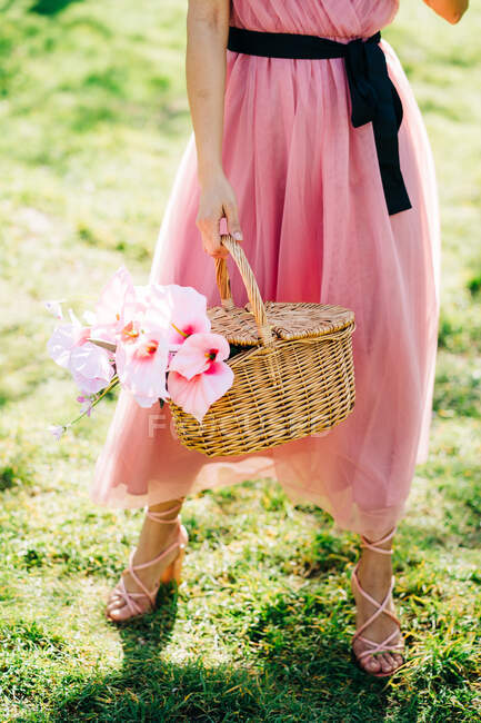 Cortada hembra irreconocible en vestido y tacones de pie con cesta con flores en un jardín - foto de stock