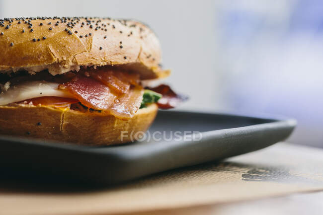 Sandwich de bagel apetitoso con tocino y pollo servido en el plato en la mesa en la cafetería - foto de stock
