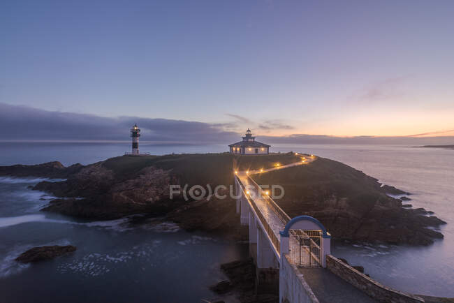Spectacular scenery of bridge leading to rocky island covered with green grass with lighthouse placed in wavy ocean in Faro Illa Pancha in Galicia in Spain in twilight — Stock Photo