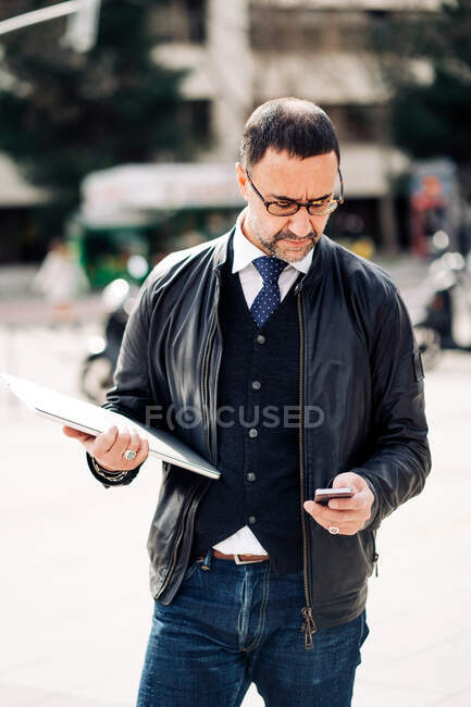 Empresario masculino de mediana edad mensajería de texto en el teléfono celular mientras está de pie en la ciudad - foto de stock