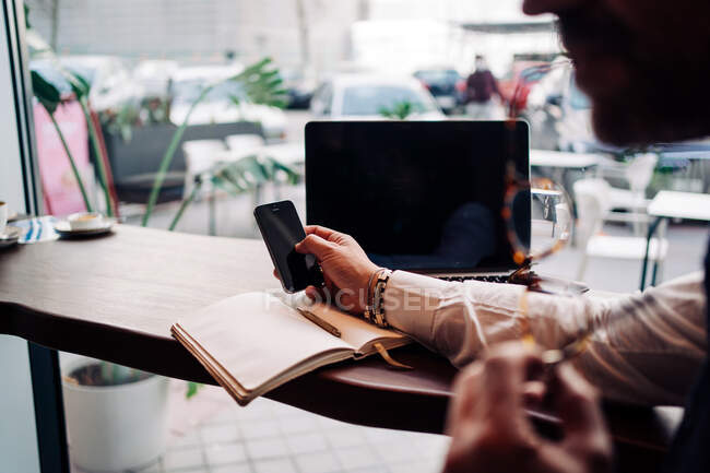 Vue latérale de la récolte anonyme homme exécutif toucher écran noir sur téléphone portable à la table avec netbook dans le café — Photo de stock