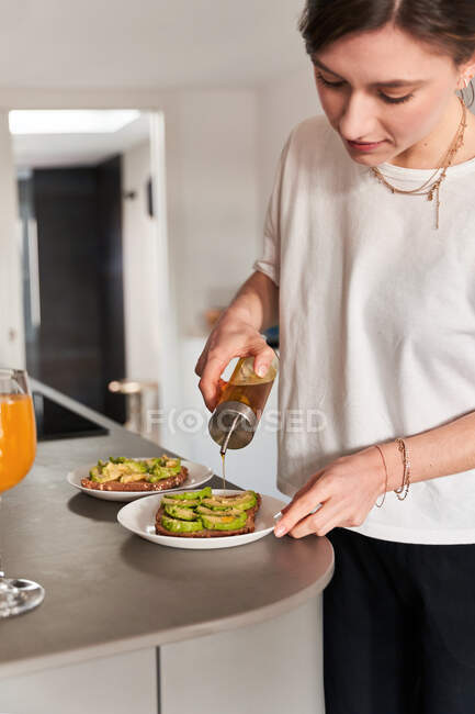 Врожай концентрував молоду жінку в повсякденному одязі, додаючи оливкову олію на смачному тості авокадо, готуючи сніданок на кухні — стокове фото