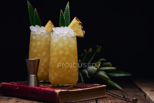 Wooden table with glasses of fresh yellow cocktails with ice cubes and pineapple pieces and leaves near spoon and shot glass placed on red book on black background — Stock Photo