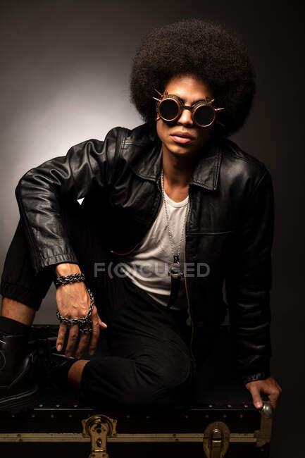 Homme afro-américain à la mode en veste et bottes en cuir assis sur fond gris — Photo de stock