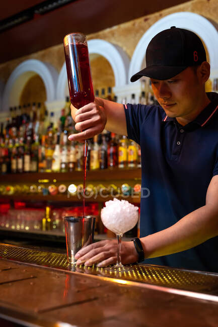 Joven camarero asiático vertiendo jugo de fresa en la coctelera mientras prepara un cóctel en el bar - foto de stock