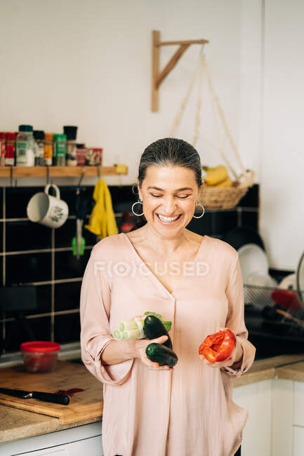 Smiling middle aged female with red bell paper leek and cucumbers looking down while standing near kitchen counter with dishware — Stock Photo