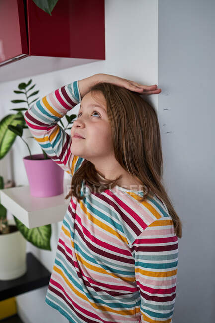 Vista lateral de la alegre linda chica de pie cerca de la pared y medir su altura y mirando hacia arriba - foto de stock