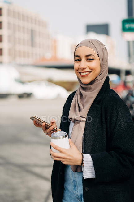 Deleitada mujer étnica en hijab y ropa de moda caminando con bebida para llevar mientras navega por Internet en un teléfono inteligente y disfruta de un fin de semana en la ciudad - foto de stock