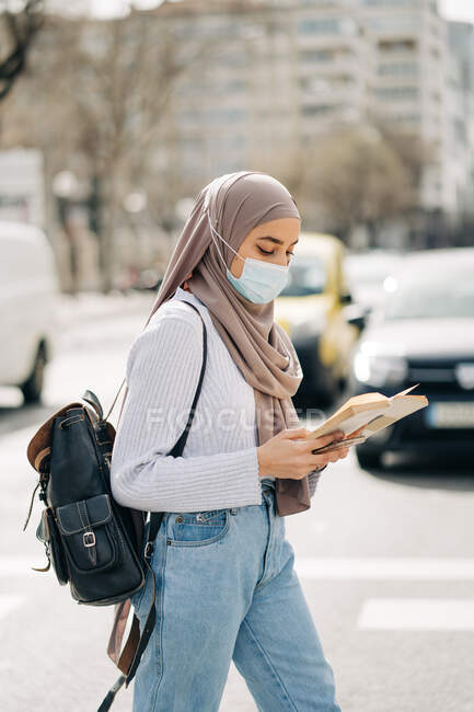 Вид збоку на етнічну жінку, що носить хустку та захисну маску, що йде по вулиці міста в сонячний день і читає книгу — стокове фото