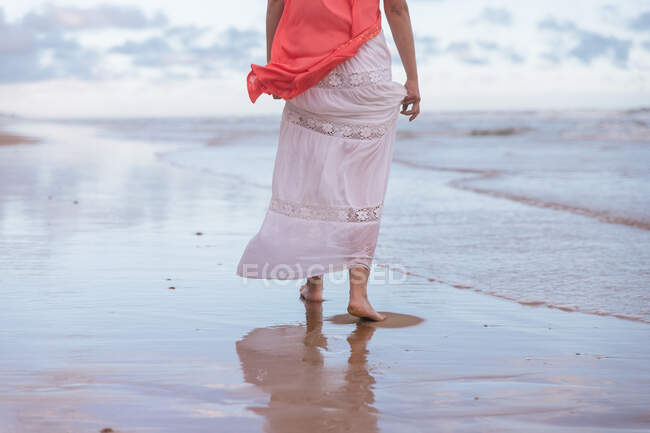 Vue arrière d'une femelle anonyme se promenant dans l'eau ondulée d'un vaste océan sur une plage de sable fin sous un ciel nuageux — Photo de stock