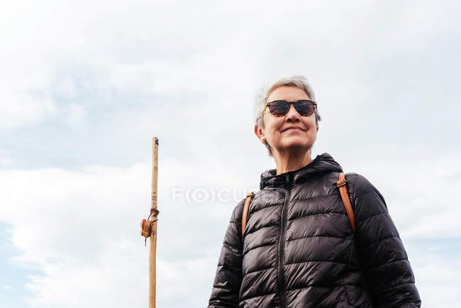 From below smiling elderly female trekker in sunglasses with gray hair looking away against cloudy sky — Stock Photo