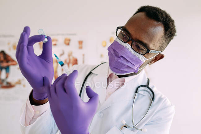 Inteligente médico afroamericano en máscara médica y guantes llenando jeringa con líquido de inyección azul en el hospital - foto de stock