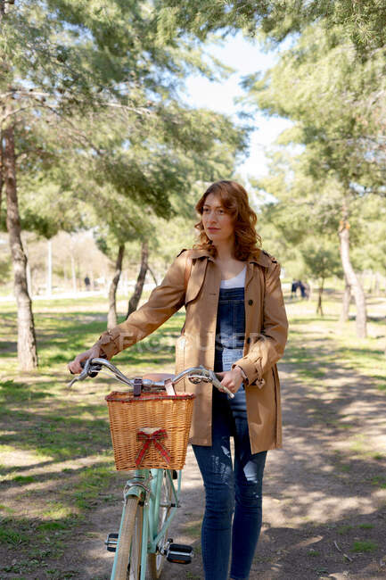 Молода жінка йде і зосереджена біля старого велосипеда з дерев'яним плетеним кошиком в парку — стокове фото