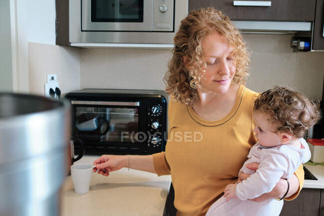 Mãe solteira alegre segurando a filha nas mãos enquanto prepara o chá na cozinha em casa durante o dia — Fotografia de Stock